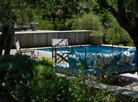 View from under the shade of mature trees to the pool and a cosy day bed with blue and white linen to the wooden terrace beyond.