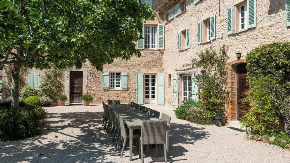 Large dining table under tree with beautiful stone-built Provencal farmhouse with green shutters in the background